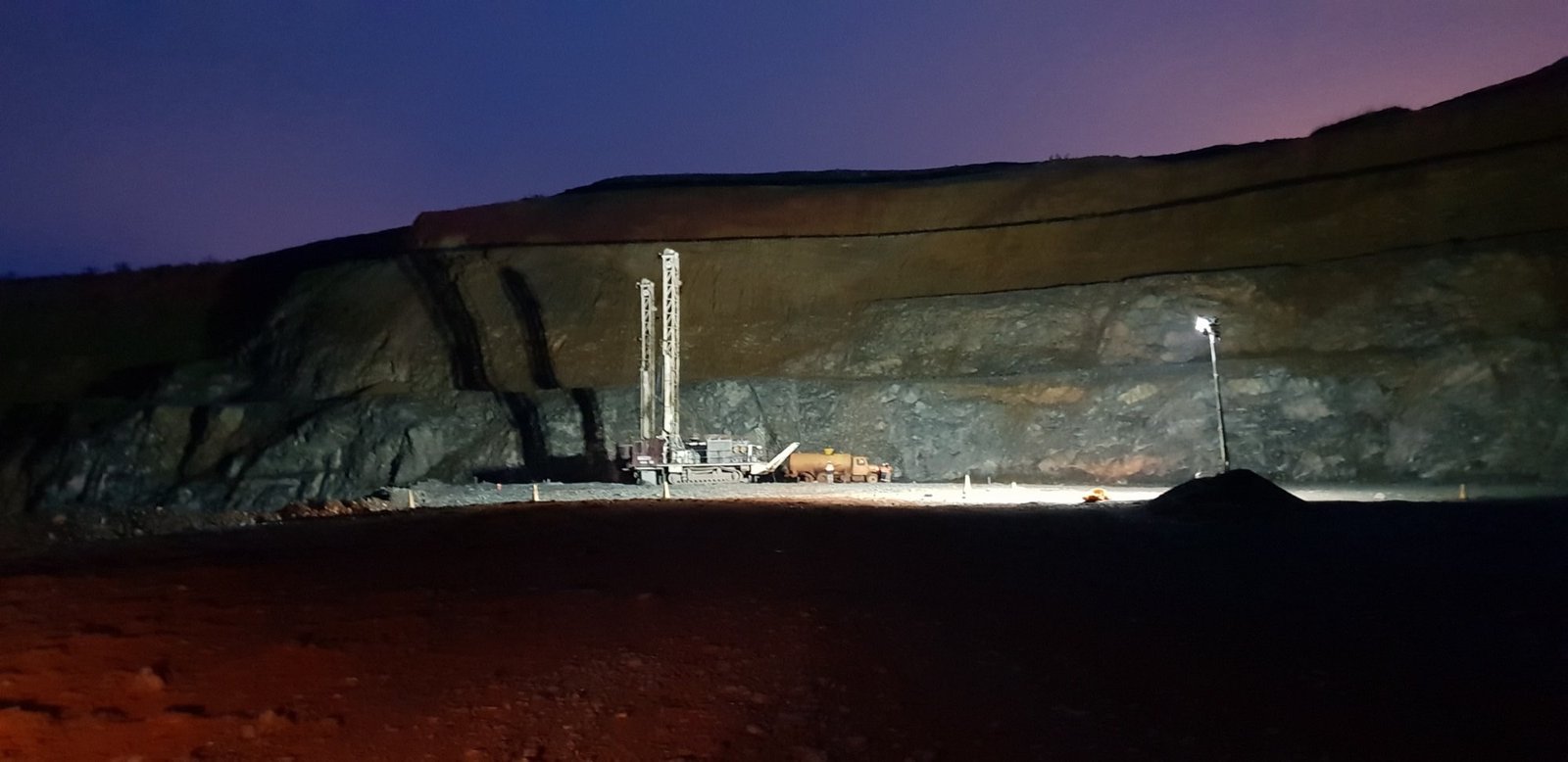 A drill rig lit by a light tower at dusk in an open pit.
